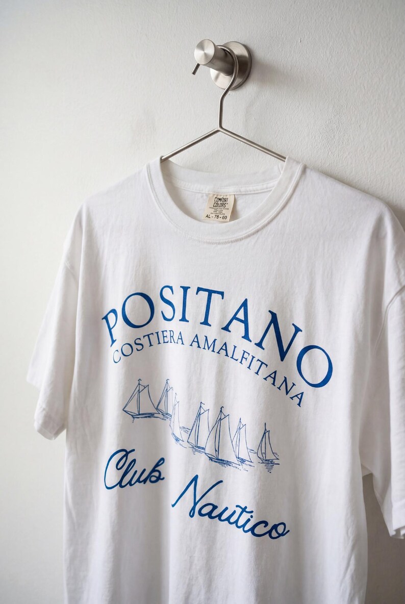 May include: A white short-sleeve t-shirt featuring blue screen-printed text that reads POSITANO COSTIERA AMALFITANA and Club Nautico, accompanied by a graphic illustration of several sailboats. The shirt has a crew neckline and a brand label.