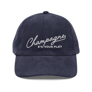 May include: Navy blue corduroy baseball cap with the word "Champagnes" in white script across the front. Below the word "Champagnes" is the phrase "S'IL VOUS PLAÎT" in white capital letters.