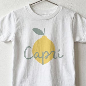 May include: A white short-sleeved t-shirt featuring a graphic of a yellow lemon with a sage green leaf and the word Capri written in sage green cursive script across the centre of the lemon. The shirt is displayed on a metal hanger.