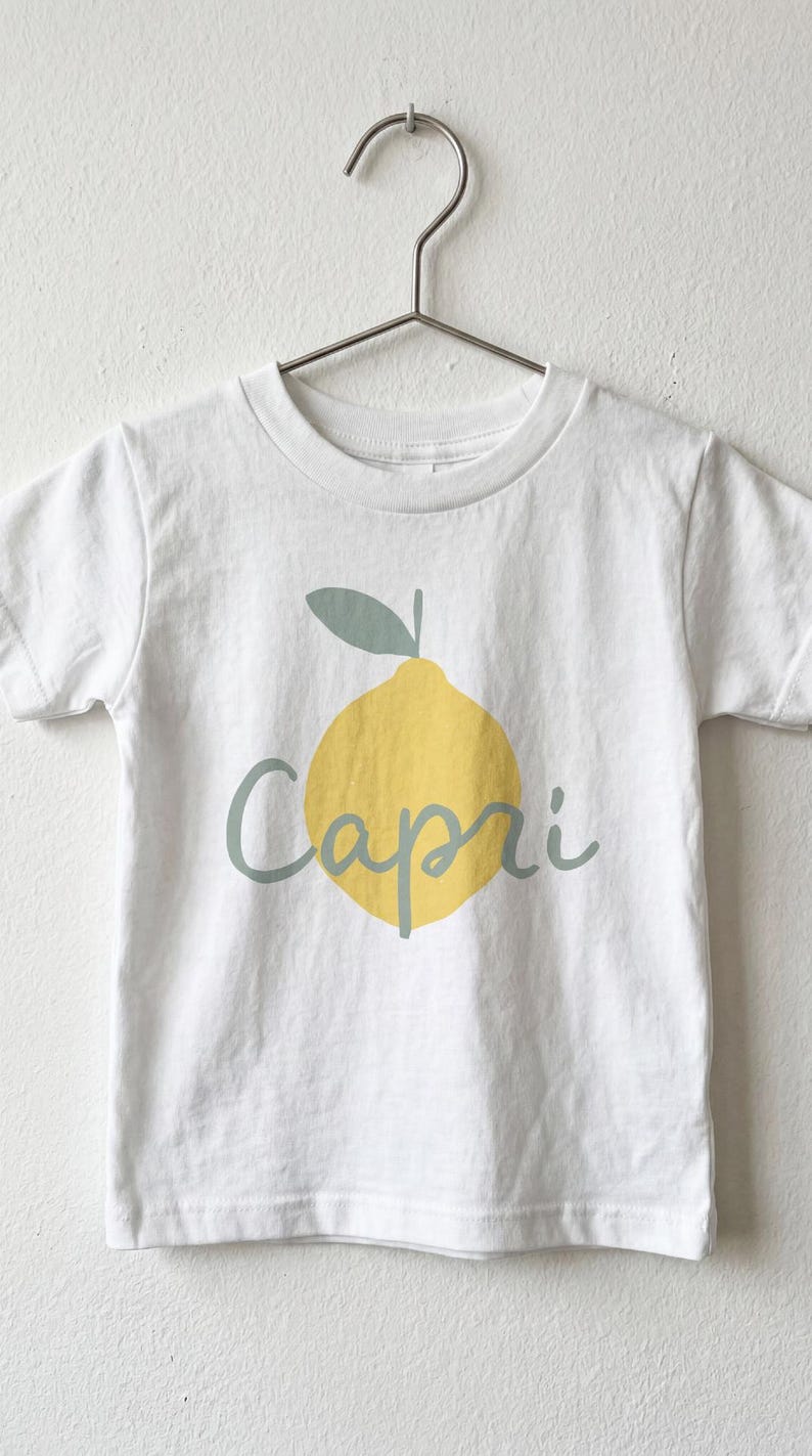 May include: A white short-sleeved t-shirt featuring a graphic of a yellow lemon with a sage green leaf and the word Capri written in sage green cursive script across the centre of the lemon. The shirt is displayed on a metal hanger.