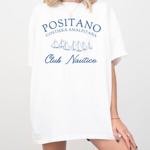 May include: A white short-sleeved t-shirt featuring blue text that reads POSITANO COSTIERA AMALFITANA and Club Nautico, accompanied by a graphic illustration of several sailboats in the center of the chest area.