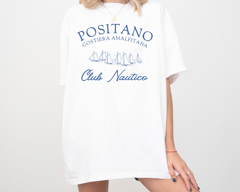 May include: A white short-sleeved t-shirt featuring blue text that reads POSITANO COSTIERA AMALFITANA and Club Nautico, accompanied by a graphic illustration of several sailboats in the center of the chest area.