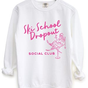 May include: White sweatshirt with the text "Ski School Dropout Social Club" in pink script. The design includes a pink illustration of a person on skis in a martini glass. The sweatshirt is hanging on a wooden hanger.