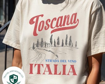 May include: Cream-colored t-shirt with the word "Toscana" in red script above a black and white illustration of a Tuscan landscape. Below the illustration, the words "Strada Del Vino" and "Italia" are printed in red.