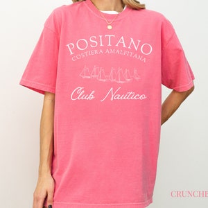 May include: A pink short-sleeved t-shirt featuring white text that reads POSITANO COSTIERA AMALFITANA and Club Nautico, along with a white line-art graphic of several sailboats. The word CRUNCHBERRY is printed in small pink letters at the bottom right.