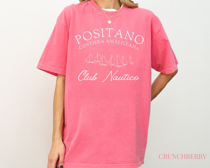 May include: A pink short-sleeved t-shirt featuring white text that reads POSITANO COSTIERA AMALFITANA and Club Nautico, along with a white line-art graphic of several sailboats. The word CRUNCHBERRY is printed in small pink letters at the bottom right.