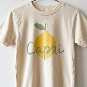 May include: A cream-coloured short-sleeve t-shirt featuring a graphic of a yellow lemon with a green leaf and the word Capri written in green cursive script across the centre. The shirt is displayed on a silver metal hanger.