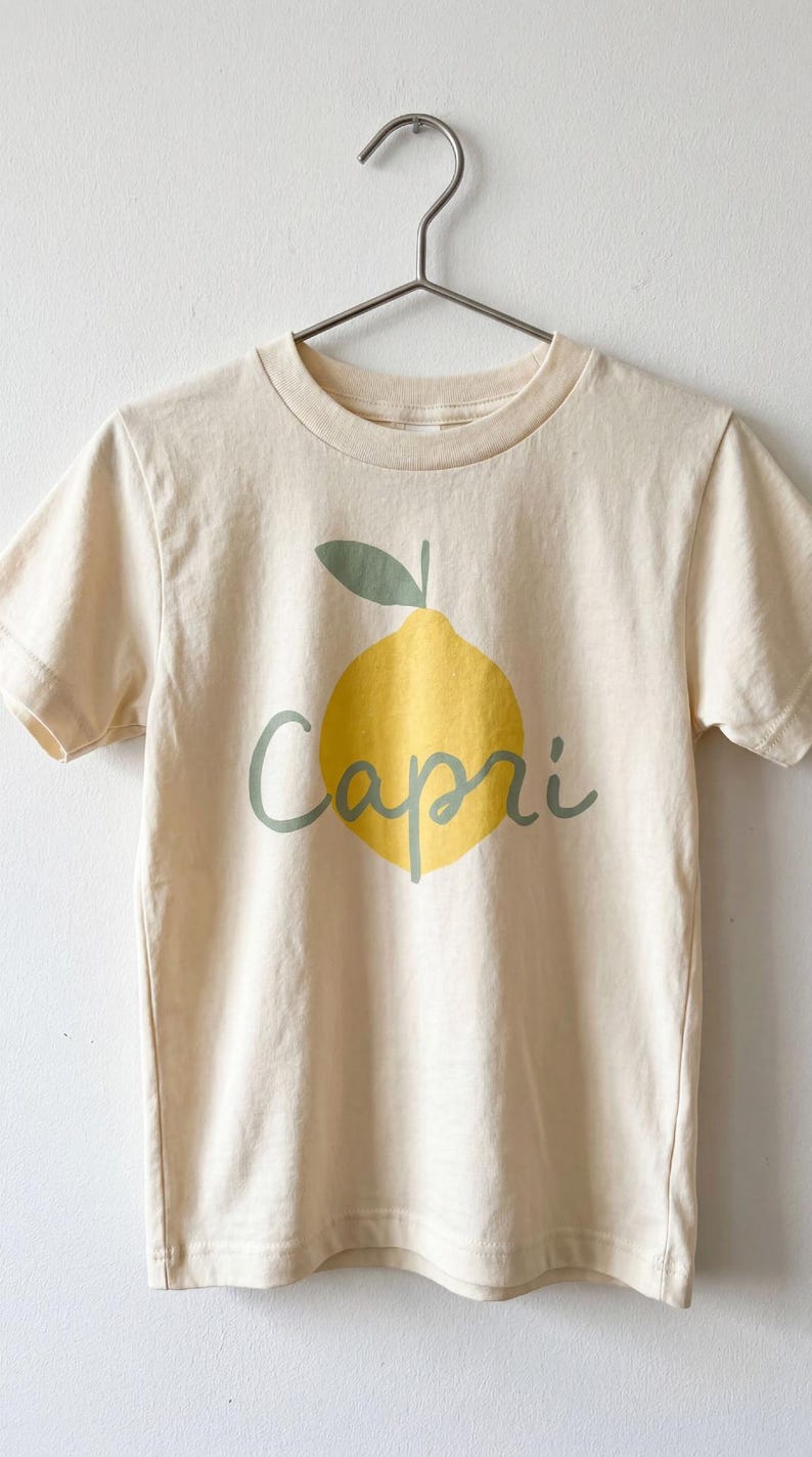 May include: A cream-coloured short-sleeve t-shirt featuring a graphic of a yellow lemon with a green leaf and the word Capri written in green cursive script across the centre. The shirt is displayed on a silver metal hanger.