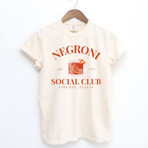 May include: A white t-shirt with a graphic design featuring a glass of Negroni with the text "Negroni dal 1919 Social Club Firenze, Italia".