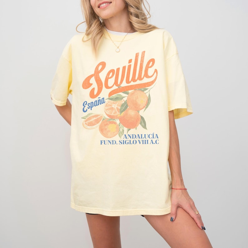 Pu&ograve; includere: Una maglietta gialla con una stampa grafica di arance e il testo "Sevilla, Espa&ntilde;a, Andaluc&iacute;a, Fund. Siglo VIII A.C."