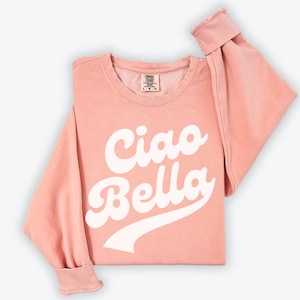 Puede incluir: Una sudadera rosa con el texto "Ciao Bella" en letras blancas en cursiva. La sudadera tiene un aspecto desgastado y un cuello redondo.