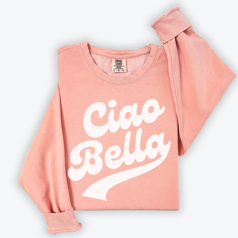 Puede incluir: Una sudadera rosa con el texto "Ciao Bella" en letras blancas en cursiva. La sudadera tiene un aspecto desgastado y un cuello redondo.