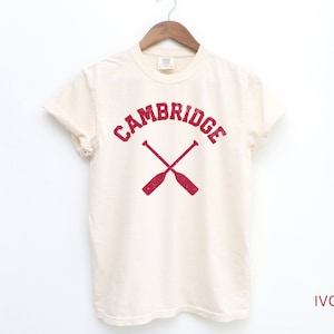 May include: A white t-shirt with a red graphic print of the word "CAMBRIDGE" in an arc with two crossed oars underneath. The text "IVORY" is printed in red at the bottom right corner of the image.
