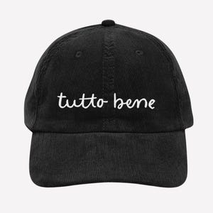May include: Black corduroy baseball cap with the white cursive text "tutto bene" embroidered on the front. The cap has a curved brim and an adjustable strap at the back. A casual accessory.