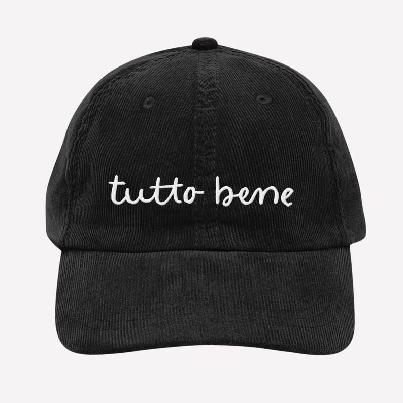 May include: Black corduroy baseball cap with the white cursive text "tutto bene" embroidered on the front. The cap has a curved brim and an adjustable strap at the back. A casual accessory.