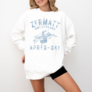 May include: White sweatshirt with a blue graphic print of a ski lift in the mountains. The text "Zermatt Switzerland" and "Apr&egrave;s-Ski" is printed on the sweatshirt.
