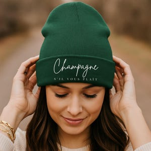 May include: A dark green beanie with the word "Champagne" and "S'il vous plaît" in white script. The beanie is made of a soft, knitted material and is worn by a person.