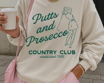 May include: Cream-colored sweatshirt with the text "Putts and Prosecco Country Club established 1985" in green script. The sweatshirt features a golf-themed graphic. The person is wearing pink and white striped shorts and a gold chain necklace.