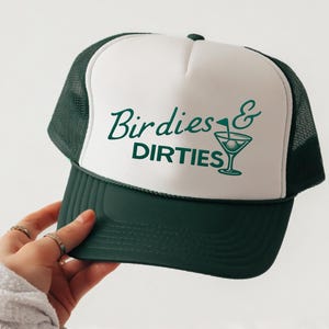 Puede incluir: Gorra de camionero verde y blanca con las palabras "Birdies & Dirties" en una fuente cursiva. La gorra presenta una copa de martini con una pelota de golf en la parte delantera. La gorra tiene una visera y una parte trasera de malla verde oscuro.