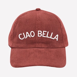 Puede incluir: Gorra de béisbol de pana rojo ladrillo con las palabras "CIAO BELLA" bordadas en blanco en la parte delantera. La gorra tiene una visera curva y un pequeño botón en la parte superior.