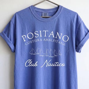 May include: A periwinkle blue short-sleeve t-shirt with white screen-printed text that reads POSITANO COSTIERA AMALFITANA and Club Nautico, featuring a graphic illustration of five sailboats in the center, hanging on a metal hanger.