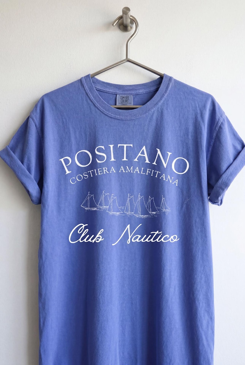 May include: A periwinkle blue short-sleeve t-shirt with white screen-printed text that reads POSITANO COSTIERA AMALFITANA and Club Nautico, featuring a graphic illustration of five sailboats in the center, hanging on a metal hanger.