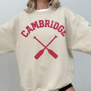 May include: A beige sweatshirt with a red vintage-style graphic that says "CAMBRIDGE" with two crossed oars underneath.