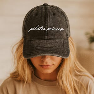 May include: A dark gray baseball cap with the words "pilates princess" embroidered in white script across the front. The cap has a curved brim and a distressed, vintage look.