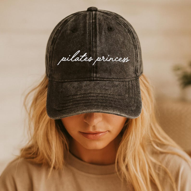 May include: A dark gray baseball cap with the words "pilates princess" embroidered in white script across the front. The cap has a curved brim and a distressed, vintage look.