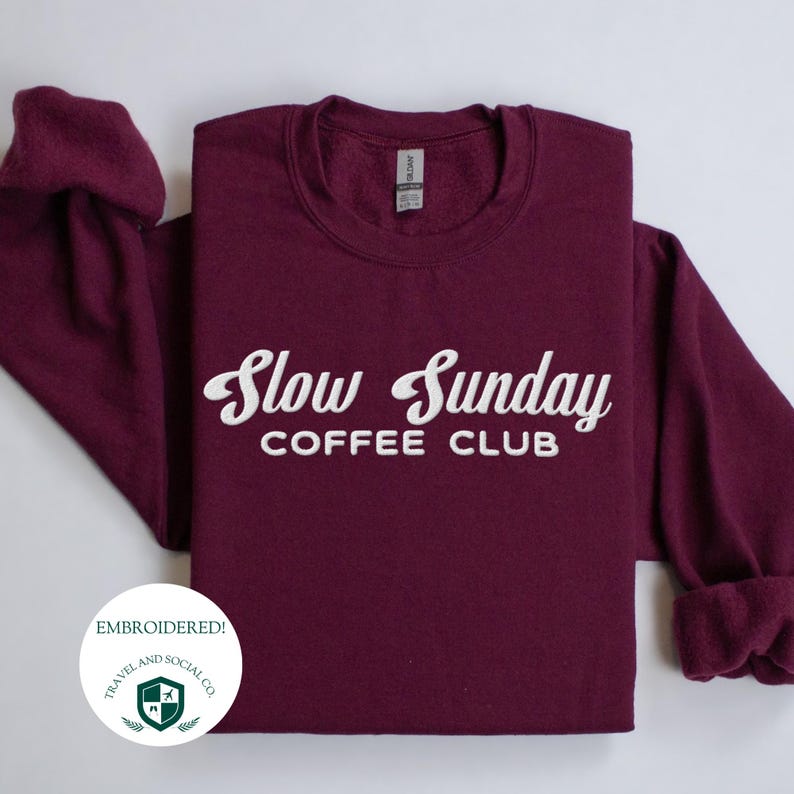 May include: Burgundy sweatshirt with white embroidered text that reads "Slow Sunday Coffee Club". The sweatshirt has a crew neck and long sleeves. The text is embroidered with a vintage style font.