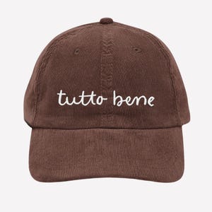 May include: A brown corduroy baseball cap with the embroidered text "tutto bene" in white. The cap has a curved brim and a top button. The hat is a solid brown color.