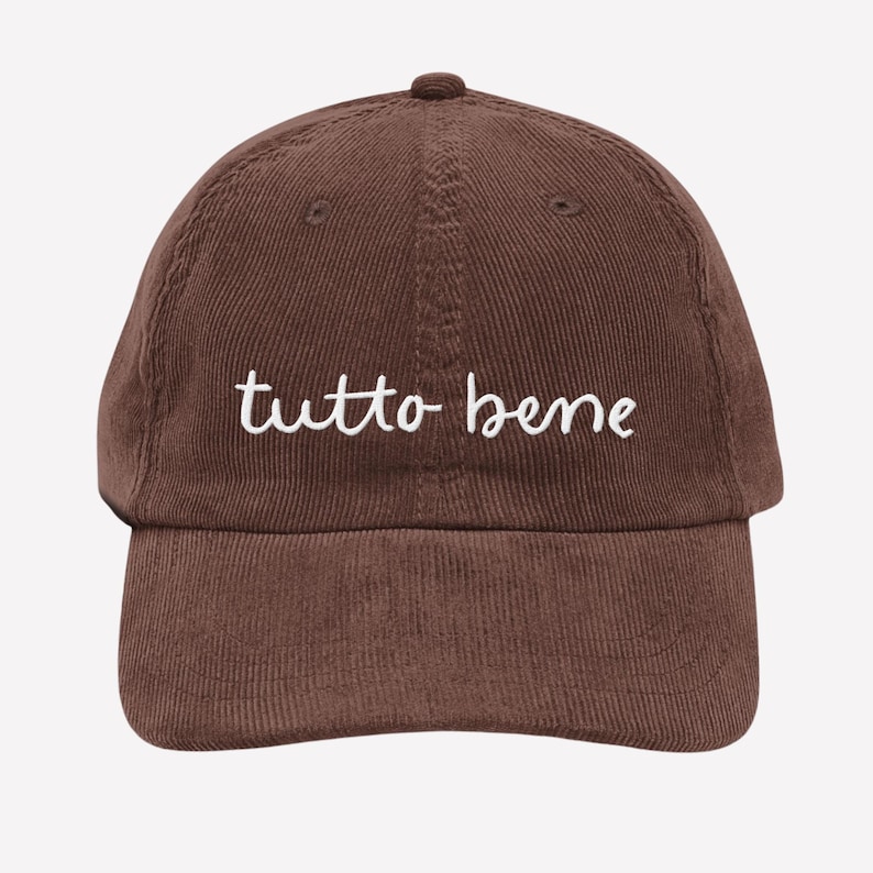 May include: A brown corduroy baseball cap with the embroidered text "tutto bene" in white. The cap has a curved brim and a top button. The hat is a solid brown color.