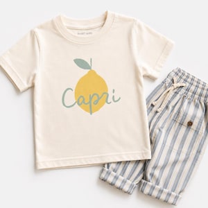 May include: A cream-coloured short-sleeve t-shirt featuring a yellow lemon graphic with a green leaf and the word Capri written in cursive script, paired with light blue and white vertically striped trousers with a drawstring waist and pockets.