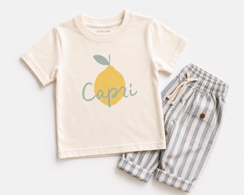 May include: A cream-coloured short-sleeve t-shirt featuring a yellow lemon graphic with a green leaf and the word Capri written in cursive script, paired with light blue and white vertically striped trousers with a drawstring waist and pockets.
