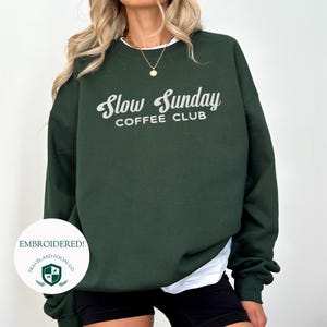 May include: A dark green sweatshirt with the text "Slow Sunday Coffee Club" embroidered in white on the front. The sweatshirt is made of a soft, comfortable material and has a relaxed fit.