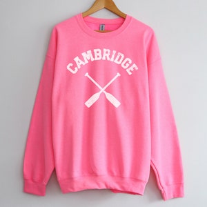 May include: A pink sweatshirt with the word "CAMBRIDGE" in white lettering and a pair of crossed oars in white.