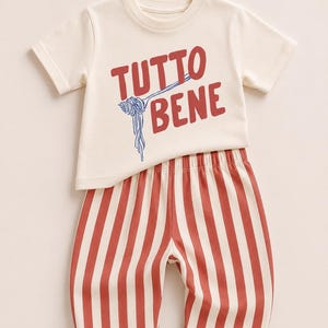 May include: A cream-colored short-sleeve t-shirt featuring the red text "TUTTO BENE" with a blue graphic of a fork twirling spaghetti, paired with shorts featuring vertical red and white stripes and an elastic waistband.