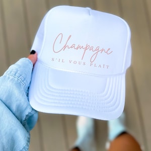 May include: White trucker hat with pink embroidered text that reads "Champagne S'il Vous Plait"