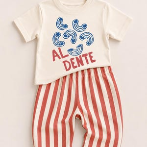 May include: A short-sleeved cream t-shirt featuring a blue macaroni pasta graphic and the red text AL DENTE, paired with pants that have vertical red and white stripes. The set consists of a crew neck top and elastic waist trousers.