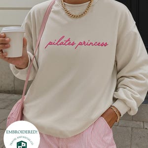 May include: Cream-colored sweatshirt with the text "pilates princess" in pink script. The sweatshirt is paired with pink and white striped shorts. A gold chain necklace and bracelet are visible. A pink purse strap is also visible.