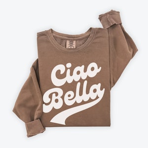 Puede incluir: Una sudadera marr&oacute;n con el texto "Ciao Bella" en letras blancas en cursiva. La sudadera tiene un corte holgado y cuello redondo.