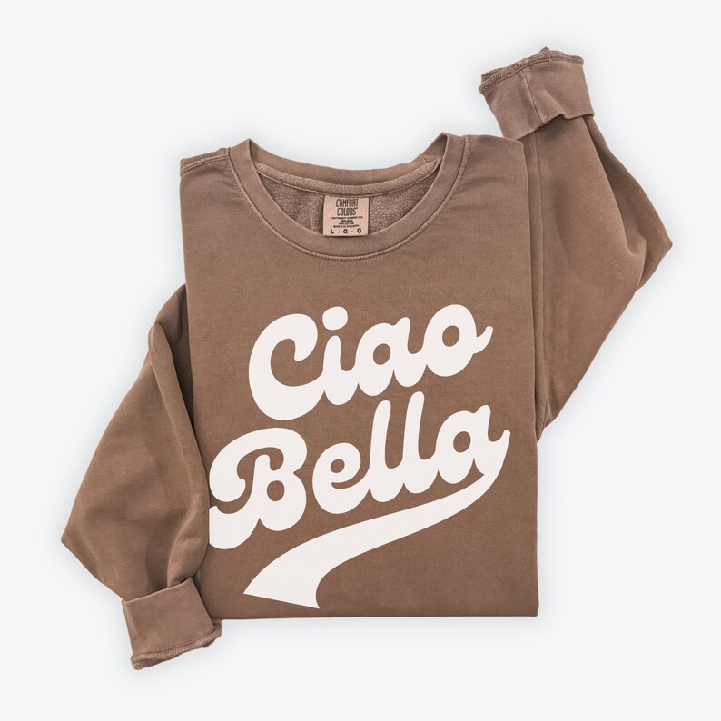 Puede incluir: Una sudadera marr&oacute;n con el texto "Ciao Bella" en letras blancas en cursiva. La sudadera tiene un corte holgado y cuello redondo.