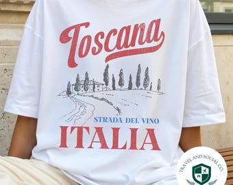 May include: White t-shirt with a red "Toscana" script and a scenic illustration of a road and buildings. Below the illustration are the words "STRADA DEL VINO ITALIA" in red. A Travel and Social Co. logo is on the lower right.