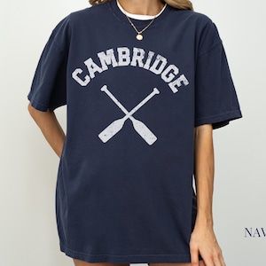 May include: A navy blue t-shirt with a white distressed graphic of the word "CAMBRIDGE" in an arched font with two crossed oars underneath.