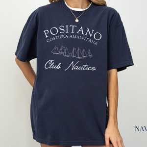 May include: A navy blue short-sleeve t-shirt featuring white screen-printed text that reads POSITANO COSTIERA AMALFITANA and Club Nautico, accompanied by a graphic illustration of several sailboats on the water in the center of the chest.