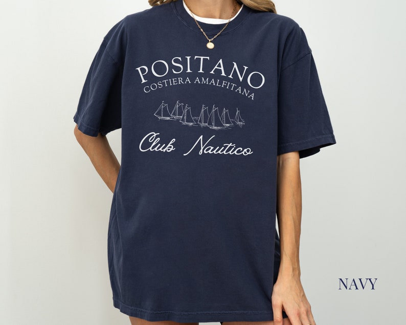 May include: A navy blue short-sleeve t-shirt featuring white screen-printed text that reads POSITANO COSTIERA AMALFITANA and Club Nautico, accompanied by a graphic illustration of several sailboats on the water in the center of the chest.