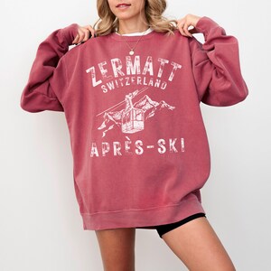 May include: A maroon sweatshirt with a white graphic print of a ski lift in the mountains. The text "Zermatt Switzerland" and "Apr&egrave;s-Ski" are printed on the sweatshirt.