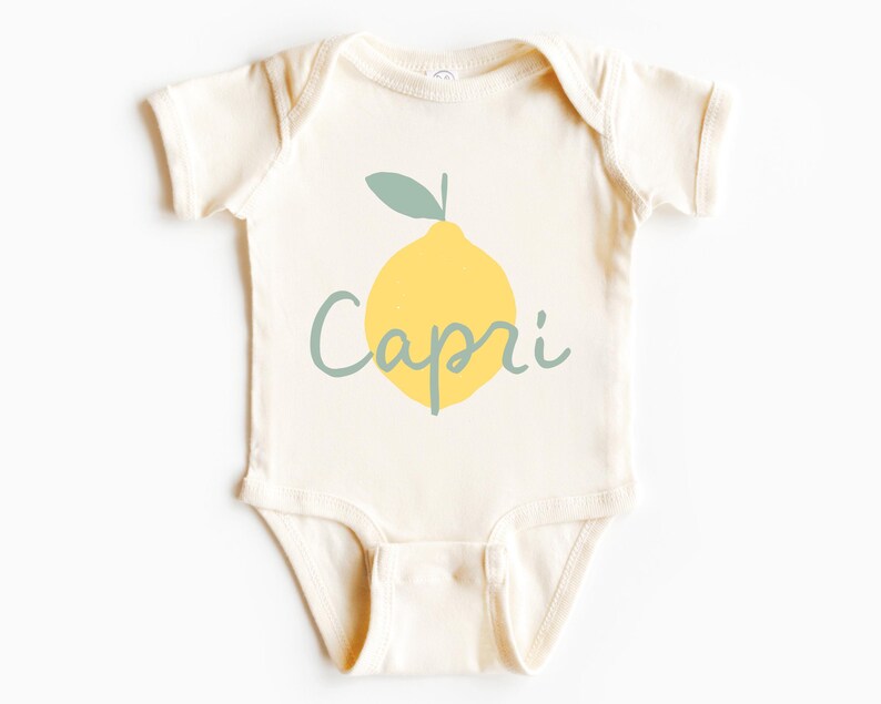 May include: A cream-coloured short-sleeve baby bodysuit featuring a graphic of a yellow lemon with a sage green leaf. The word Capri is printed across the centre of the lemon in a sage green cursive script font, centred on the chest area.