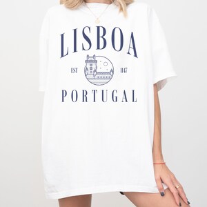 Puede incluir: Camiseta blanca con un dise&ntilde;o gr&aacute;fico azul que presenta una imagen estilizada de un edificio, una luna y estrellas. El texto "LISBOA EST 1147 PORTUGAL" est&aacute; impreso en la camiseta.