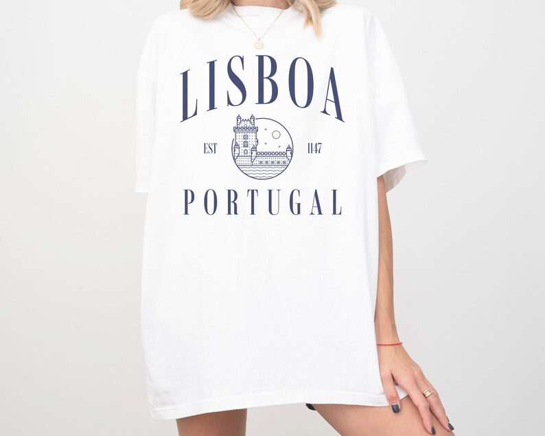 Puede incluir: Camiseta blanca con un dise&ntilde;o gr&aacute;fico azul que presenta una imagen estilizada de un edificio, una luna y estrellas. El texto "LISBOA EST 1147 PORTUGAL" est&aacute; impreso en la camiseta.
